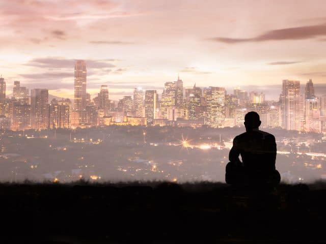 A picture of a man sitting on top of a hill and he is looking lonely while looking at the city.