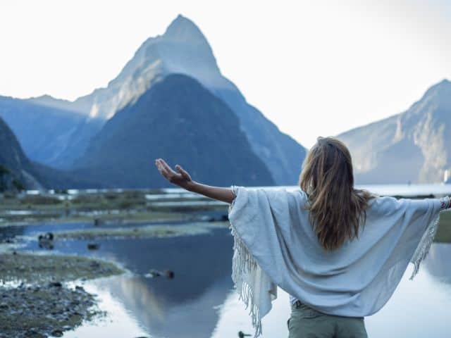 A picture of a woman standing with her arms outstretched in front of the Mitre Peak mountain in Milford Sound. New Zealand.