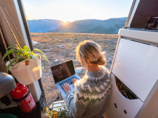 A picture of a woman working remotely on her laptop while sitting in a van on top of a mountain while trying slow travel.