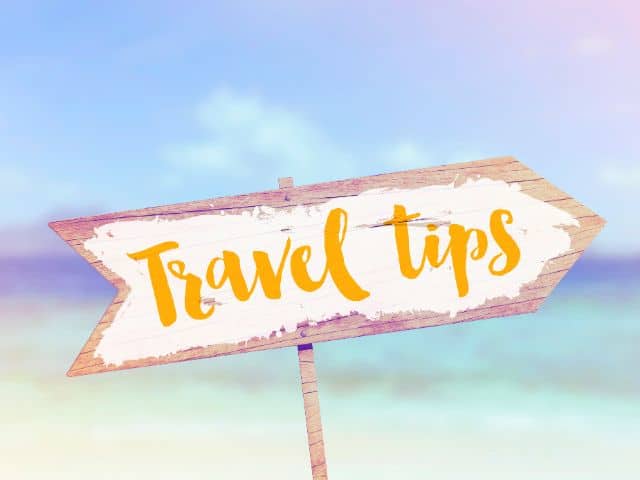 A picture of an arrow-shaped wooden sign painted in white with the words "travel tips" written on it and the background is a beach.