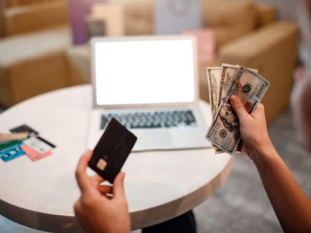 A picture of a person holding a black credit card in their left hand and money in their right hand with a laptop in front of them.