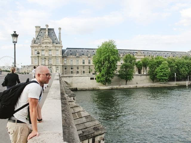 A picture of a male tourist wearing a bag while outside of the Louvre Museum in Paris, France.