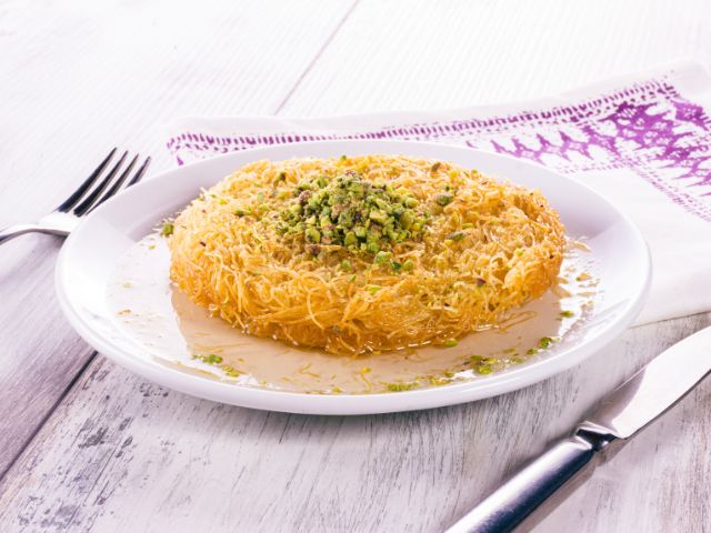 A picture of a piece of Kunafa placed in a white plate with a knife and a fork next to it.